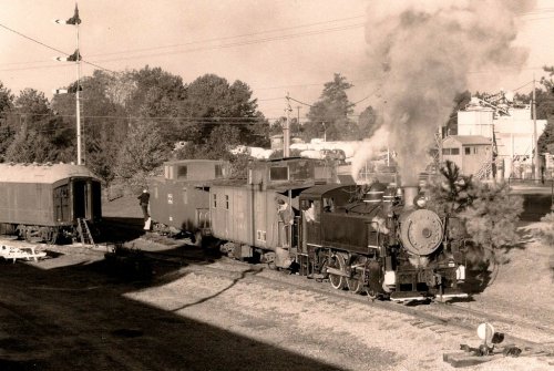 A vintage steam locomotive with a tender and caboose, releasing steam, surrounded by trees and railway signals.