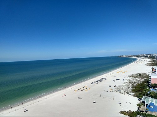 A sunny beach with clear blue water, white sand, and scattered beach chairs under a clear sky.