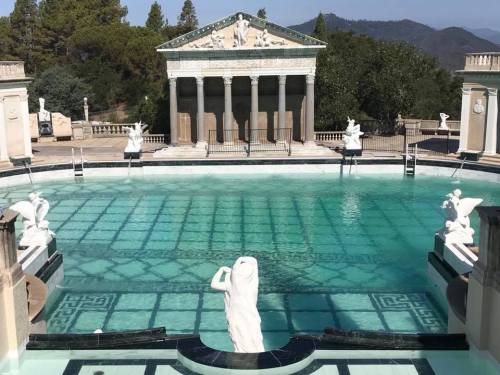 A grand pool surrounded by classical architecture and sculptures, set against a mountainous backdrop.