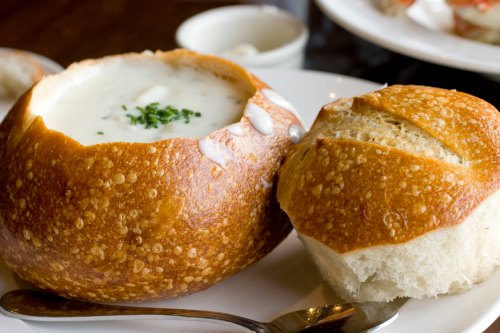 A bowl of creamy soup served in a bread bowl, accompanied by a small piece of crusty bread on a plate.