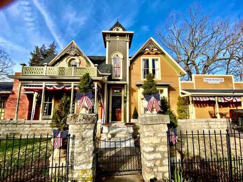 A charming historic house with American flags, surrounded by a stone fence and greenery, under a clear blue sky.