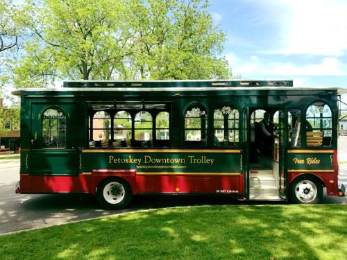 A green and red trolley with "Petoskey Downtown Trolley" signage parked on a grassy area under trees.
