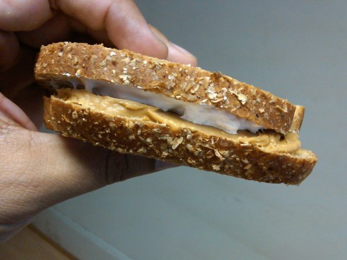 A hand holding a sandwich made with oat bread, filled with peanut butter and a layer of white cream.