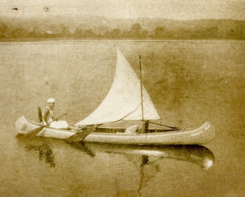 A person in a canoe with a sail, paddling on calm water, surrounded by a serene landscape.
