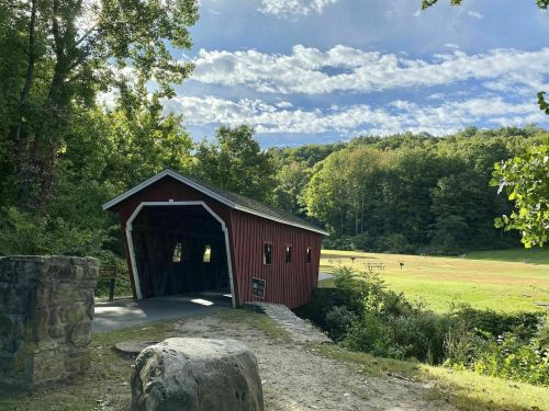 A red covered bridge surrounded by trees and a grassy field under a blue sky with scattered clouds.