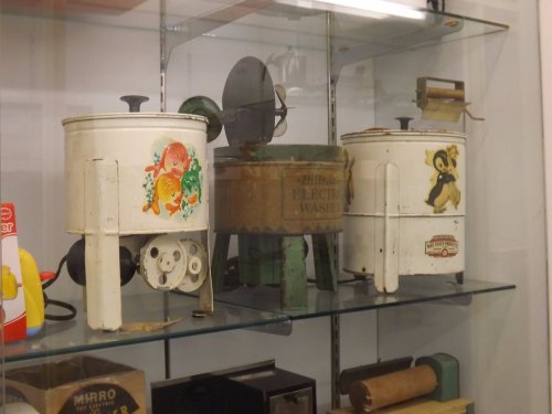 Three vintage washing machines displayed in a glass case, featuring colorful designs and retro branding.