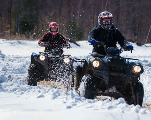 Two riders on ATVs kick up snow while navigating a winter landscape.