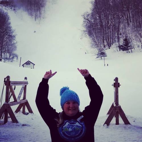 A person in a blue hat stands in the snow, smiling and pointing upwards at a snowy slope in a winter landscape.