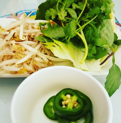 A plate of fresh herbs and bean sprouts, with a small bowl of sliced green chili peppers.