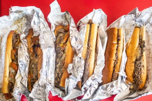 A row of six sandwiches wrapped in foil, filled with meat, peppers, and onions, on a red surface.