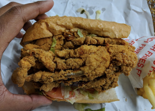 A hand holding a large fried chicken sandwich with lettuce, tomato, and a toasted bun, served with fries in the background.