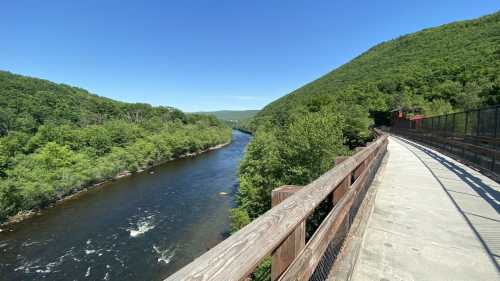 A scenic view of a river winding through lush green hills, with a wooden walkway alongside. Clear blue sky above.