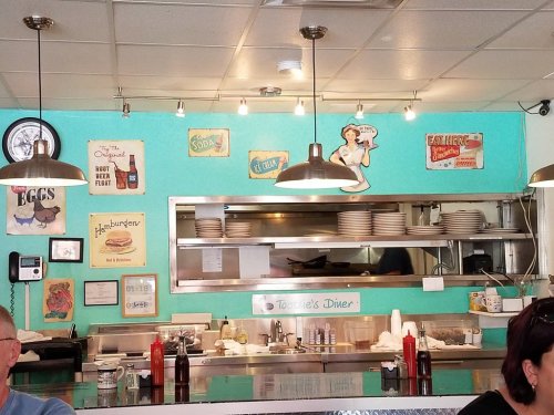 A retro diner interior with teal walls, vintage signs, and a visible kitchen area. Tables and condiments are in the foreground.