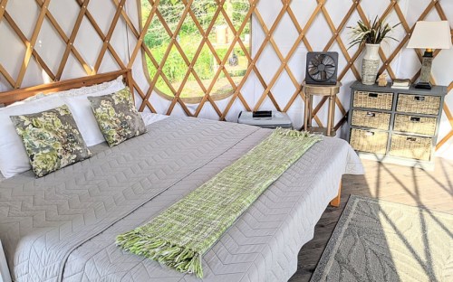 Cozy yurt interior with a bed, decorative pillows, a fan, and a nightstand, surrounded by wooden lattice walls.