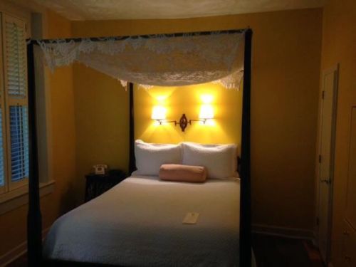 Cozy bedroom with a four-poster bed, soft lighting, and yellow walls, featuring a decorative canopy and a nightstand.