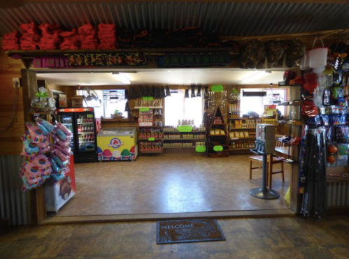 Interior of a small store with shelves stocked with snacks, drinks, and various merchandise. A welcome mat is visible.