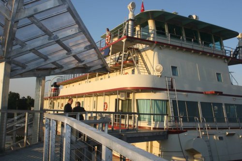 A view of a large boat docked at a pier, with people on the deck and a clear sky in the background.