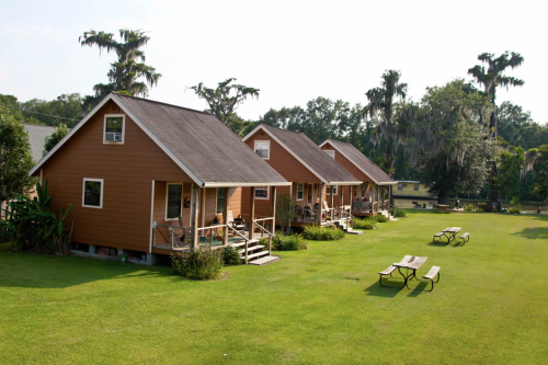 Three cozy cabins with porches sit on a grassy area, surrounded by trees and picnic tables under a clear sky.