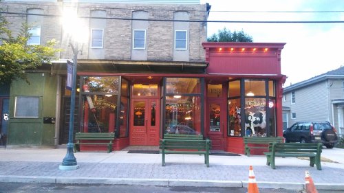 A charming storefront with large windows, red and green accents, and benches outside, set in a quaint town.