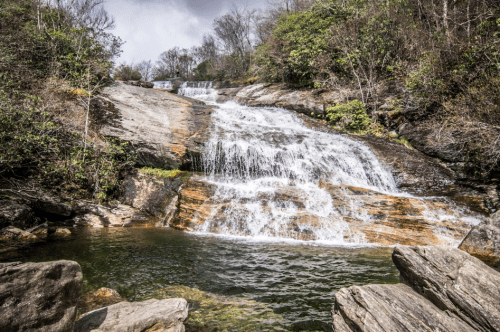 A cascading waterfall flows over rocky terrain into a serene pool, surrounded by lush greenery and a cloudy sky.