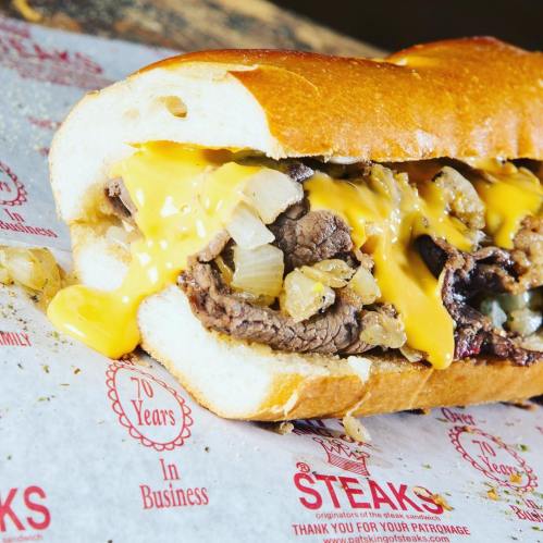 Close-up of a cheesesteak sandwich with melted cheese and onions on a toasted hoagie roll, resting on branded paper.