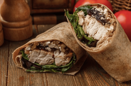 A close-up of a sliced wrap filled with chicken, greens, nuts, and cranberries, resting on a wooden surface.