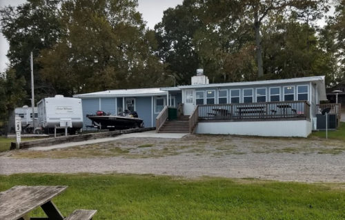 A blue building with a deck, parked boat, and RVs nearby, surrounded by trees and gravel.