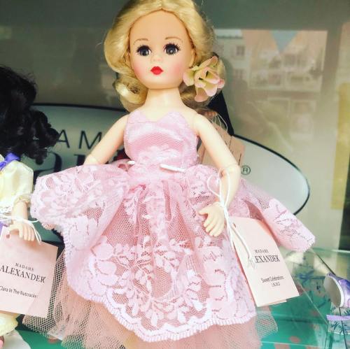 A vintage Madame Alexander doll in a pink lace dress with a flower in her hair, displayed in a showcase.