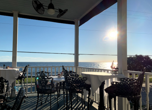 A seaside restaurant patio with empty tables, overlooking a calm ocean and bright sun in a clear blue sky.