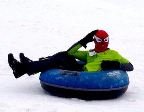 A person in a Spider-Man mask and green jacket relaxes on a blue snow tube in a snowy landscape.