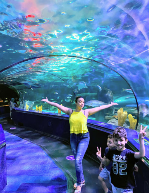 A woman in a yellow top poses with arms outstretched in an aquarium, while two children smile and make peace signs.