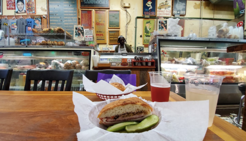 A sandwich on a plate with pickles, with a counter and a person in the background at a deli. Drinks are on the table.