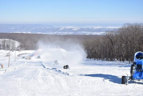 Snow-covered landscape with snowmaking machines and a ski slope in the background, under a clear blue sky.