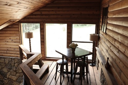 Cozy wooden interior with a table and stools, large windows, and a staircase leading down. Natural light fills the space.