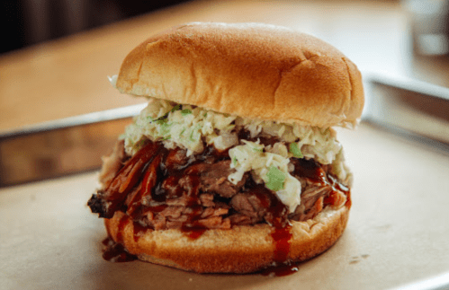 A close-up of a pulled pork sandwich topped with coleslaw and barbecue sauce, served on a soft bun.