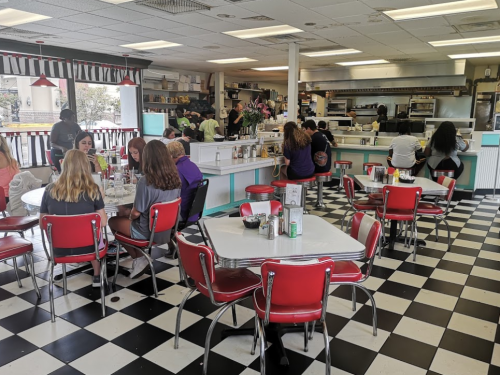 A retro diner with checkered floors, red chairs, and patrons enjoying meals at tables. The kitchen is visible in the background.