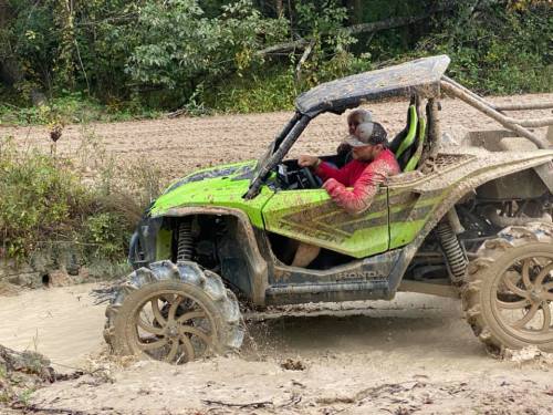 A muddy off-road vehicle drives through a puddle, with two people inside enjoying the ride.