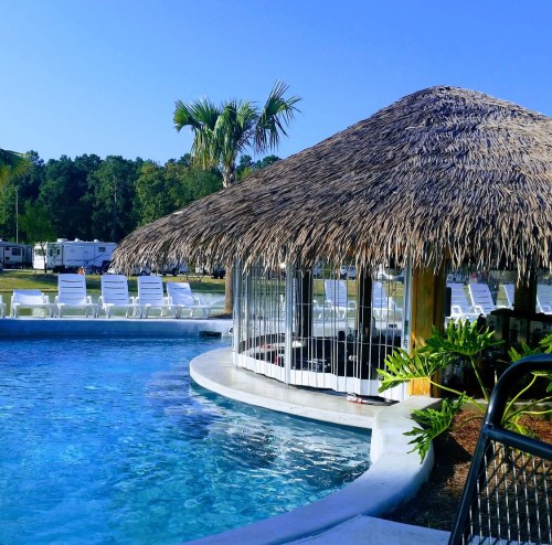 A tropical pool area with a thatched roof cabana, lounge chairs, and palm trees under a clear blue sky.