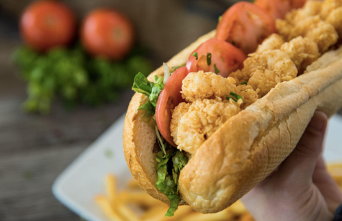 A hand holding a large sandwich filled with shrimp, lettuce, and tomato, with fries and tomatoes in the background.