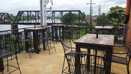 Outdoor seating area with wooden tables and black chairs overlooking a river and a train bridge in the background.
