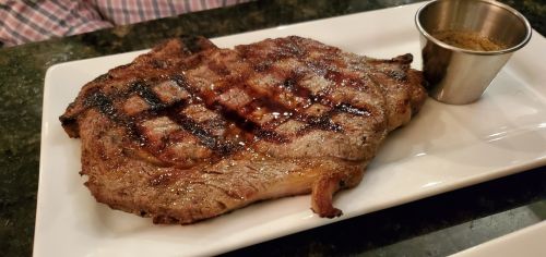 A grilled steak on a white plate, served with a small cup of seasoning on the side.