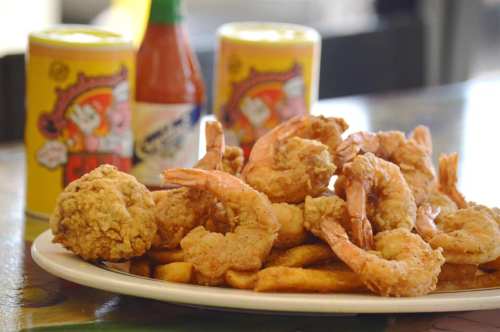 A plate of fried shrimp and chicken, served with fries, hot sauce, and drinks in the background.