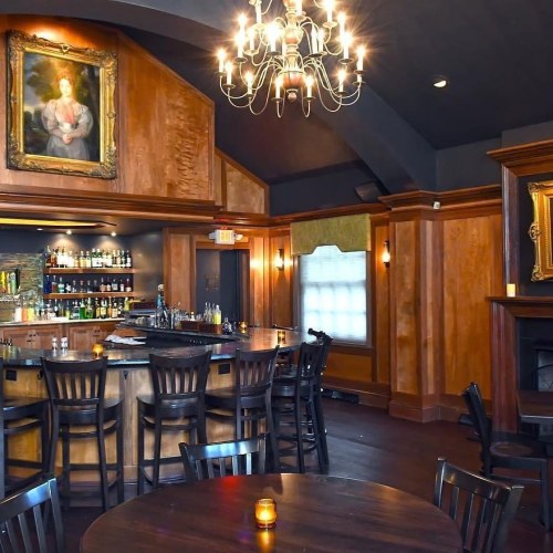 A cozy bar area with wooden decor, a chandelier, and a large portrait on the wall, featuring empty tables and chairs.