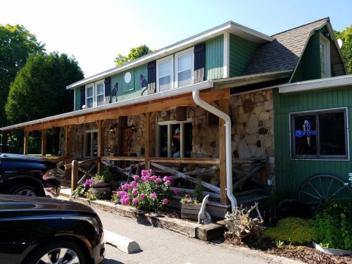 A green and stone building with a porch, flower beds, and a neon sign, surrounded by parked cars and trees.