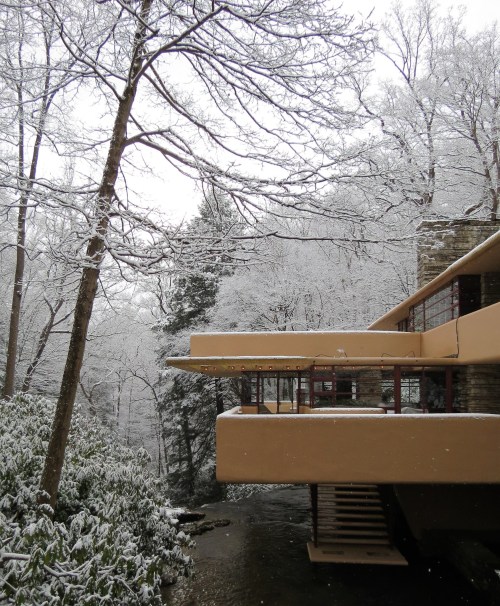 A modern house with a cantilevered design surrounded by snow-covered trees and a stream.