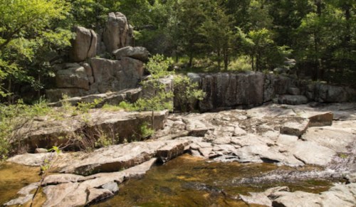 Rocky landscape with greenery and a small stream flowing through, surrounded by trees and boulders.