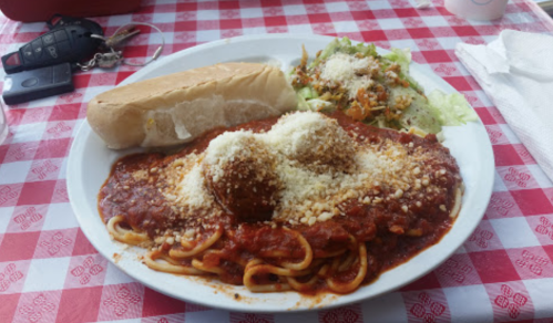 A plate of spaghetti with meatballs and sauce, topped with cheese, served with a side salad and a bread roll on a checkered tablecloth.
