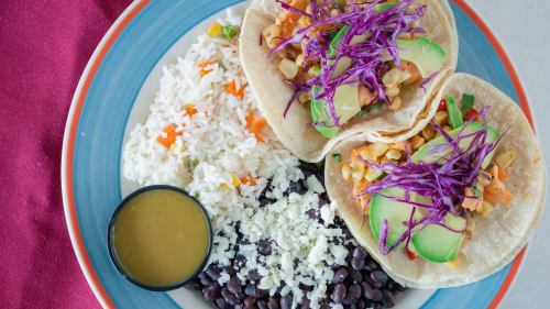 A colorful plate featuring two tacos with cabbage, rice, black beans, and a small cup of sauce.