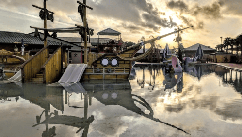 A colorful pirate ship playground reflected in a calm pool, with a cloudy sky in the background.