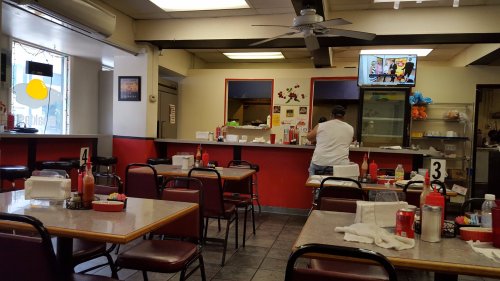 A casual diner interior with empty tables, ketchup bottles, and a chef working behind the counter. A TV is on the wall.